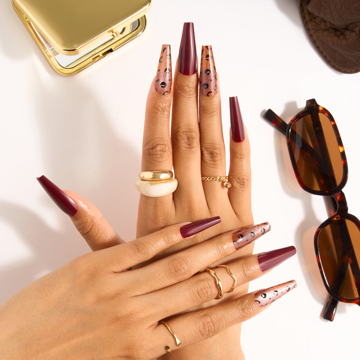 Thumbnail: L.A. Colors Glam Mani Nail Tips in So Feisty (CNT774) worn on both hands: long burgundy coffin nails with rose-gold leopard-print accent nails, styled with gold rings and tortoiseshell sunglasses.