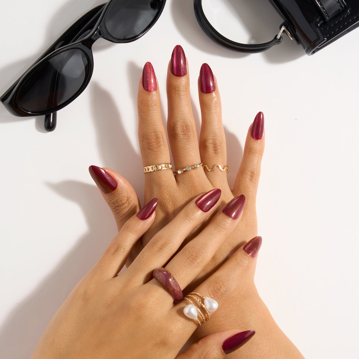 L.A. Colors Glam Mani Nail Tips in Ruby (CNT775) worn on both hands: shimmering deep red almond nails, styled with gold rings, a pearl ring and black sunglasses.
