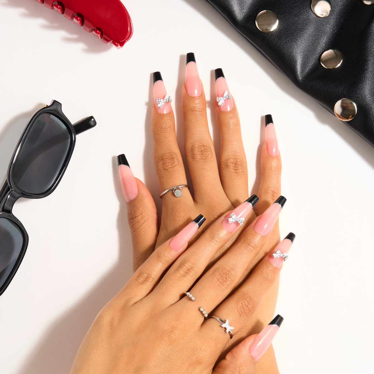 L.A. Colors Glam Mani Nail Tips in French Twist (CNT776) worn on both hands: long coffin nails with black French tips and silver bow gem accents, styled with a black studded bag and black sunglasses.
